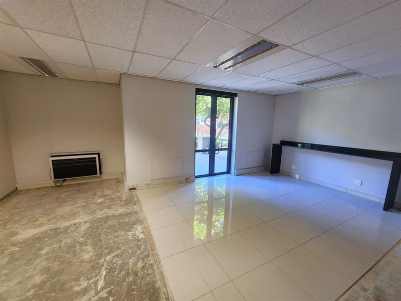 An empty office space featuring a suspended ceiling, recessed lighting, wall-mounted air conditioning, and a mix of tiled and unfinished flooring with a built-in counter.