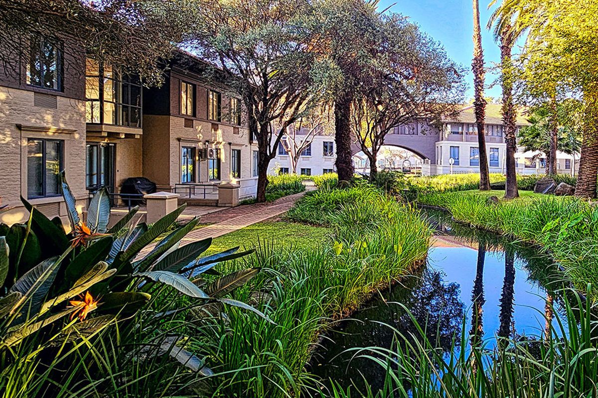 A scenic view of the office complex exterior featuring a landscaped garden, a tranquil water feature, and paved walkways between building wings.