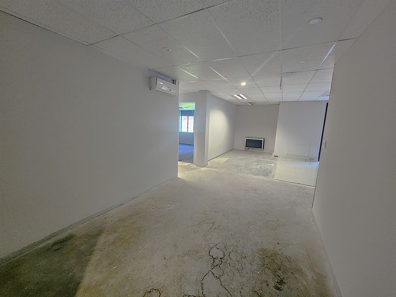 An empty office space featuring a suspended ceiling, recessed lighting, and a wall-mounted air conditioning unit, currently requiring flooring repairs and finishing.