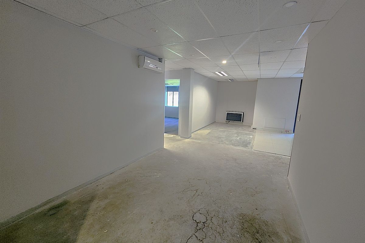 An empty office space featuring a suspended ceiling, recessed lighting, and a wall-mounted air conditioning unit, currently requiring flooring repairs and finishing.