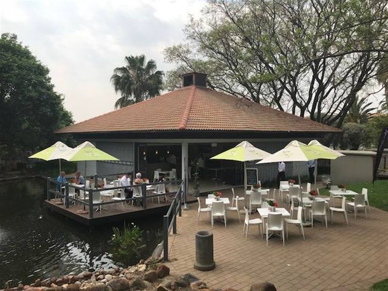 An outdoor entertainment area featuring a covered structure with a tiled roof, adjacent timber decking over a pond, and a paved patio space.