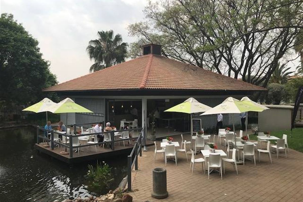 An outdoor entertainment area featuring a covered structure with a tiled roof, adjacent timber decking over a pond, and a paved patio space.