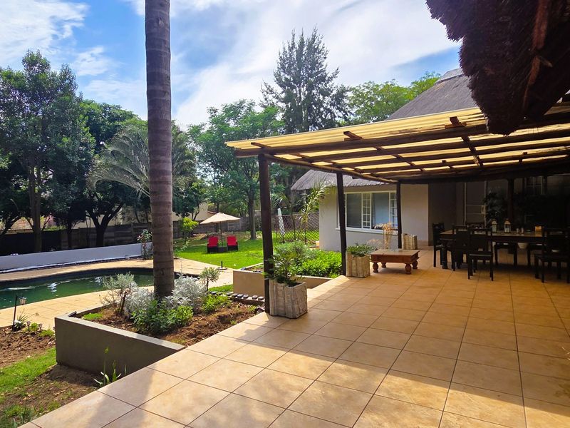 Spacious covered patio featuring tiled flooring and a timber pergola, overlooking a well-maintained garden and swimming pool area.