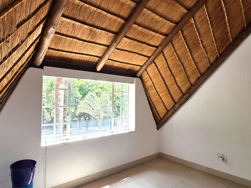 An empty bedroom featuring a distinctive vaulted thatched roof with exposed timber rafters, tiled flooring, and a large window overlooking the garden.