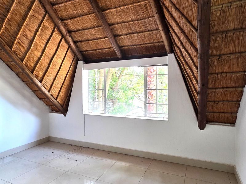 An empty bedroom featuring a vaulted thatched ceiling with exposed timber rafters, tiled flooring, and a large window overlooking greenery.