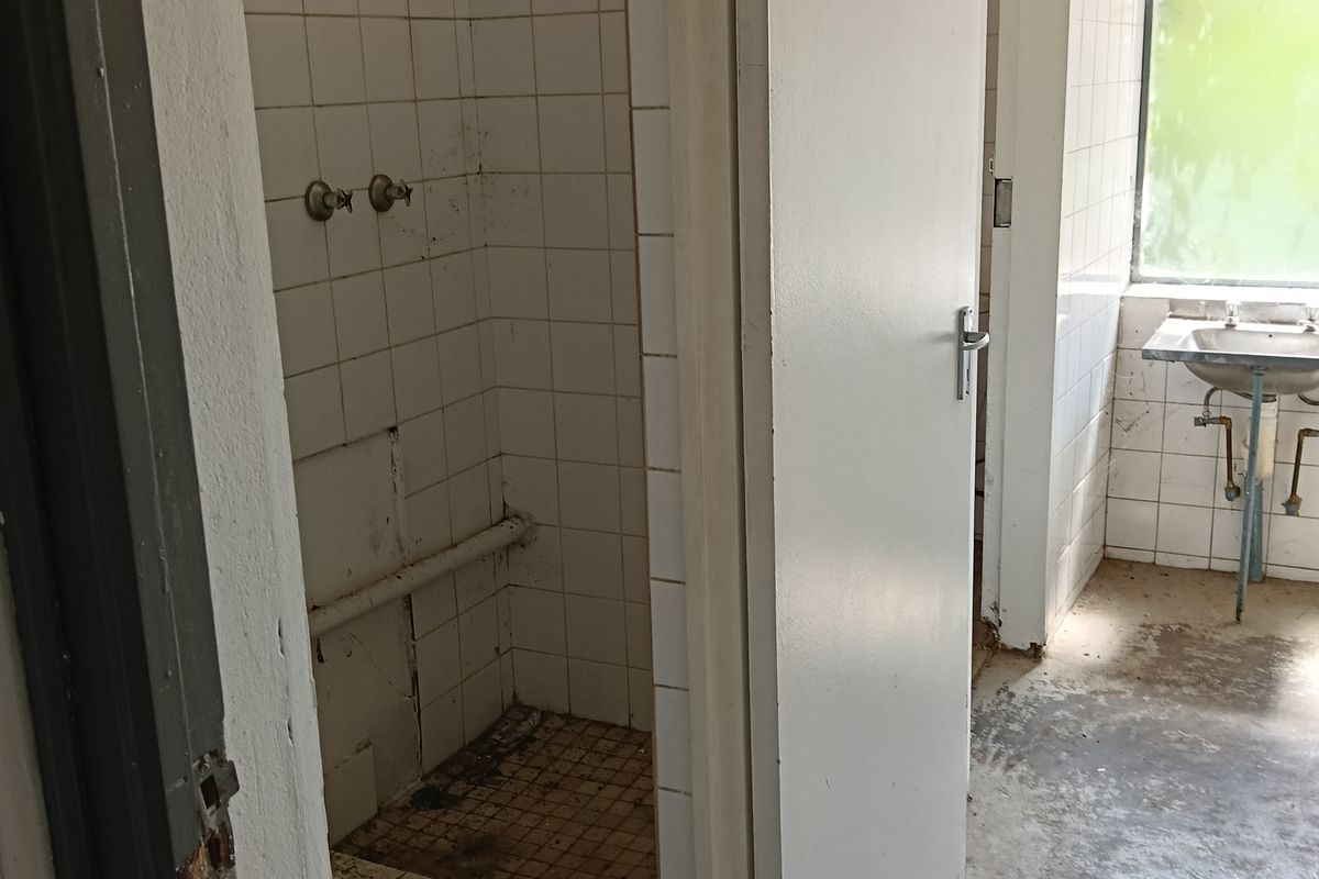 A dilapidated industrial bathroom facility featuring a tiled shower stall, wall-mounted basin, and exposed plumbing, requiring significant renovation and repair.