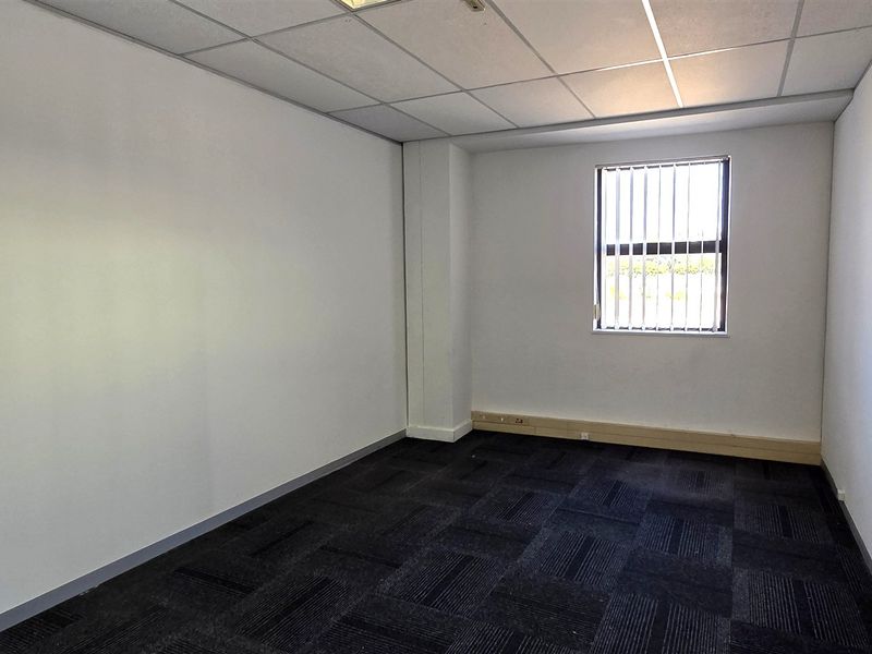 A vacant office space featuring dark carpet tiles, a suspended ceiling, and a window with security bars, offering a clean and functional workspace.