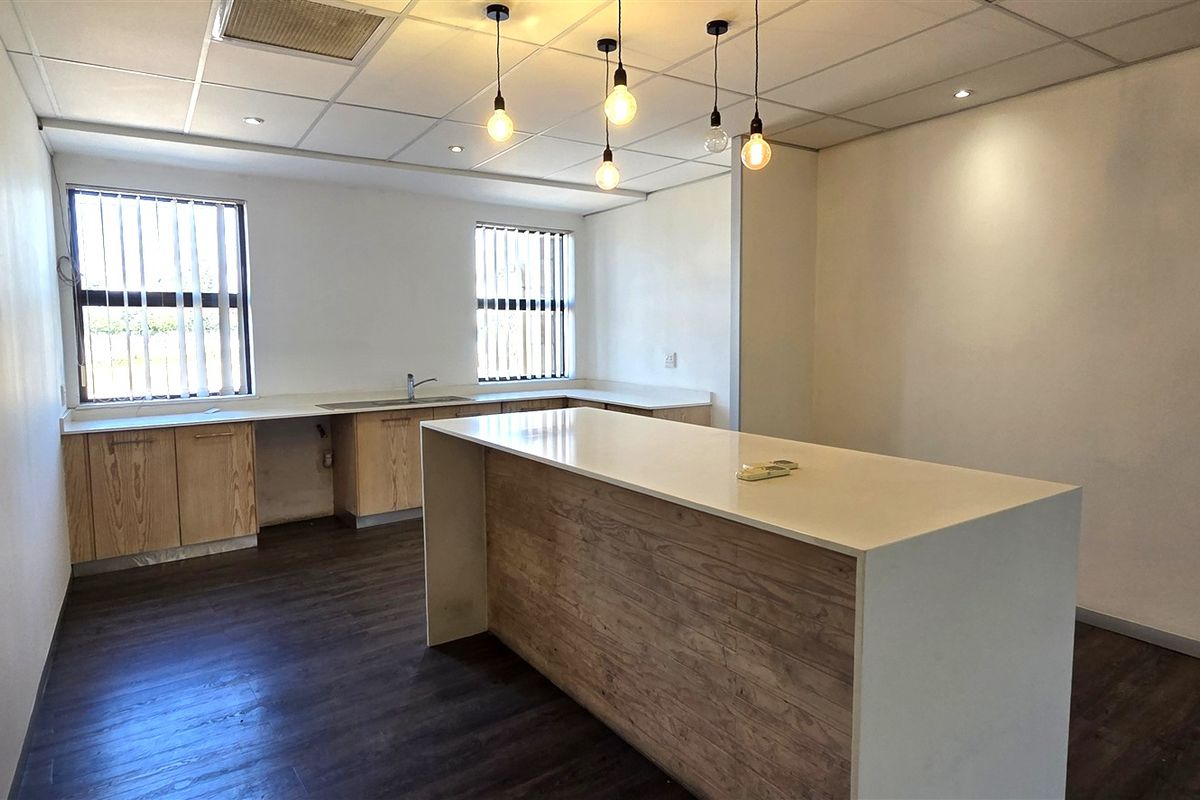A clean, modern office kitchenette featuring a central island with stone countertop, wood-finish cabinetry, and contemporary pendant lighting in an open-plan workspace.