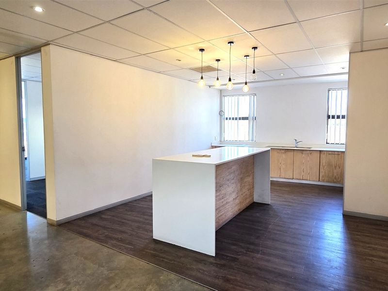 A modern, open-plan office space featuring a kitchenette with built-in cabinetry, a central island, suspended ceilings, and a mix of concrete and laminate flooring.