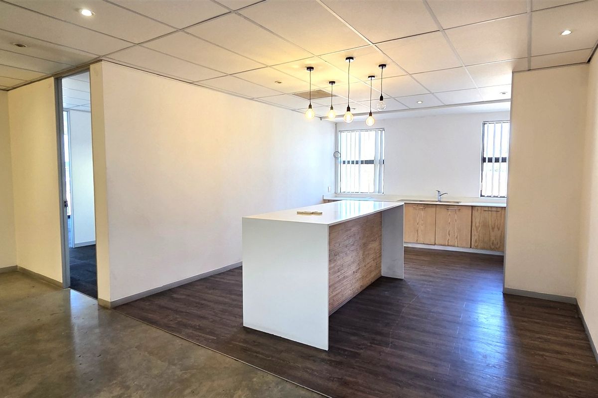 A modern, open-plan office space featuring a kitchenette with built-in cabinetry, a central island, suspended ceilings, and a mix of concrete and laminate flooring.