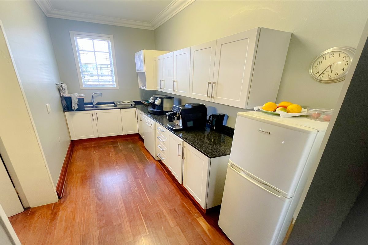 A functional office kitchen featuring white cabinetry, granite countertops, a stainless steel sink, and hardwood flooring.
