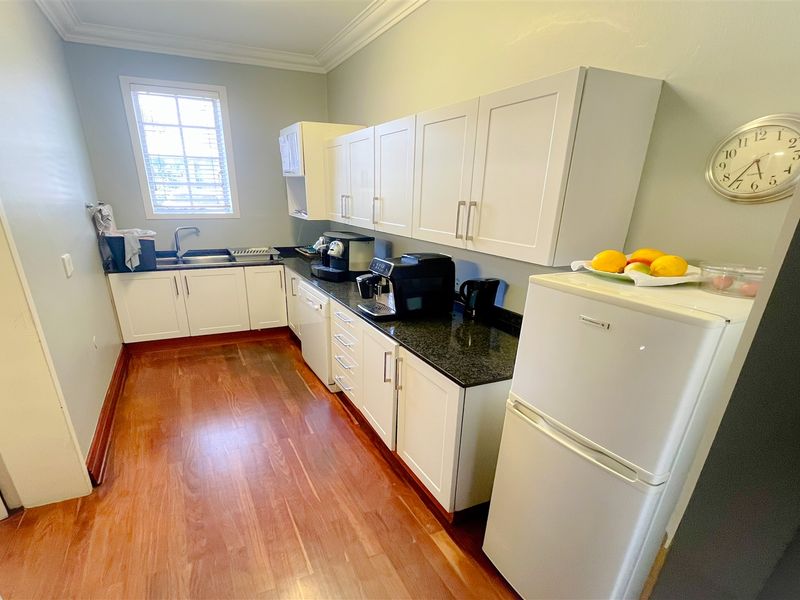 A functional office kitchen featuring white cabinetry, granite countertops, a stainless steel sink, and hardwood flooring.