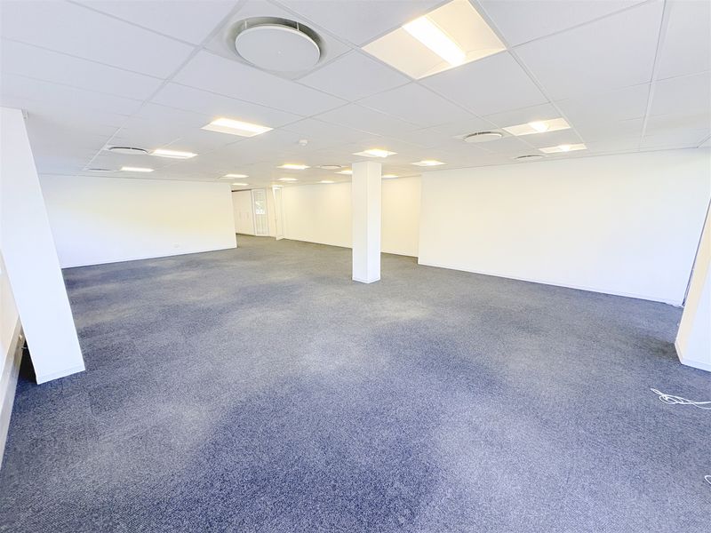 A spacious, open-plan office area featuring a suspended ceiling with recessed lighting, carpeted flooring, and structural support columns.