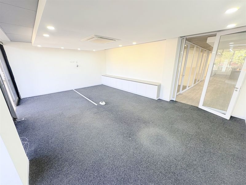 A bright, modern office space featuring carpet flooring, recessed ceiling lighting, integrated air conditioning, and a glass partition wall with built-in storage cabinetry.