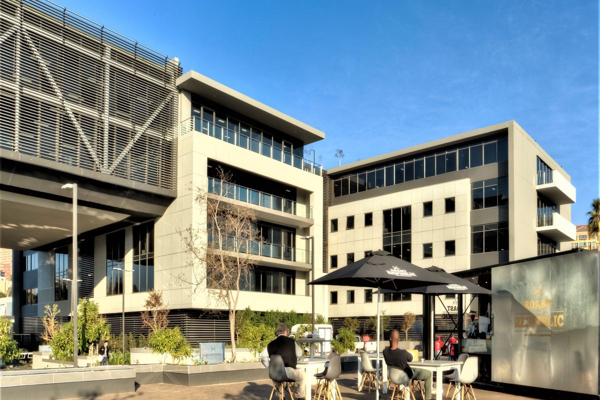 A modern office building exterior featuring a paved courtyard area with contemporary architectural design, large glass windows, and integrated sun louvres.