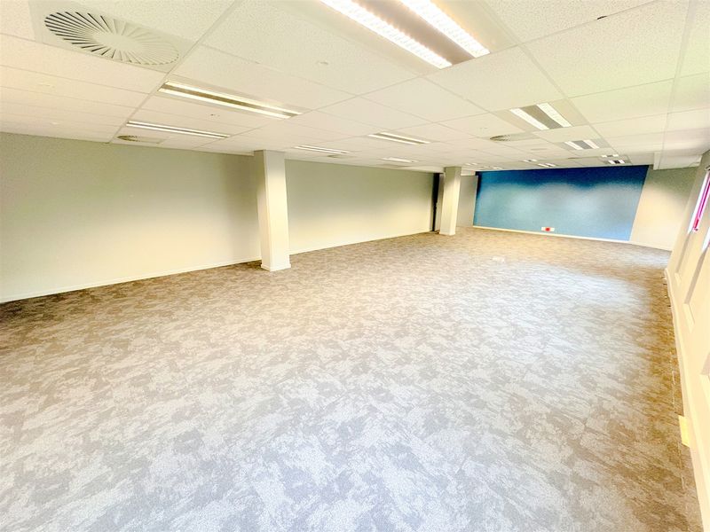 A spacious, open-plan office area featuring carpet tiles, a suspended ceiling with recessed lighting, and structural columns, providing a clean and versatile workspace.