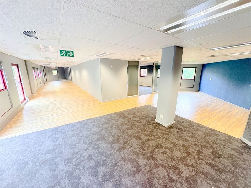 Spacious open-plan office area featuring a mix of laminate and carpet flooring, suspended ceilings with recessed lighting, and a central structural column.