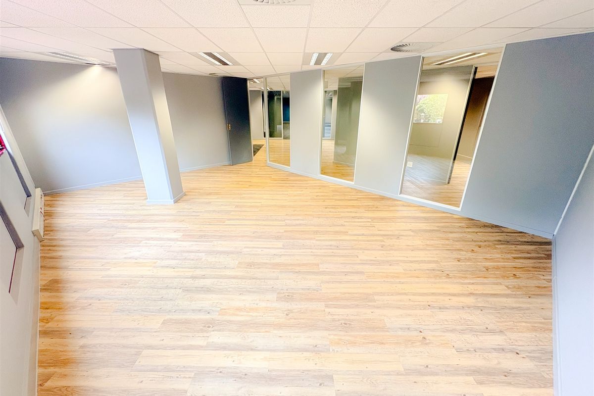 A spacious, modern open-plan office area featuring light wood-look laminate flooring, glass partitioning, and a clean, neutral aesthetic suitable for professional use.