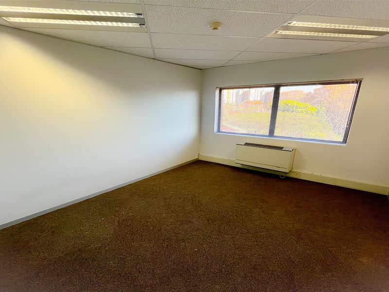 A vacant office space featuring a suspended ceiling with recessed lighting, carpeted flooring, and a window with integrated HVAC unit.