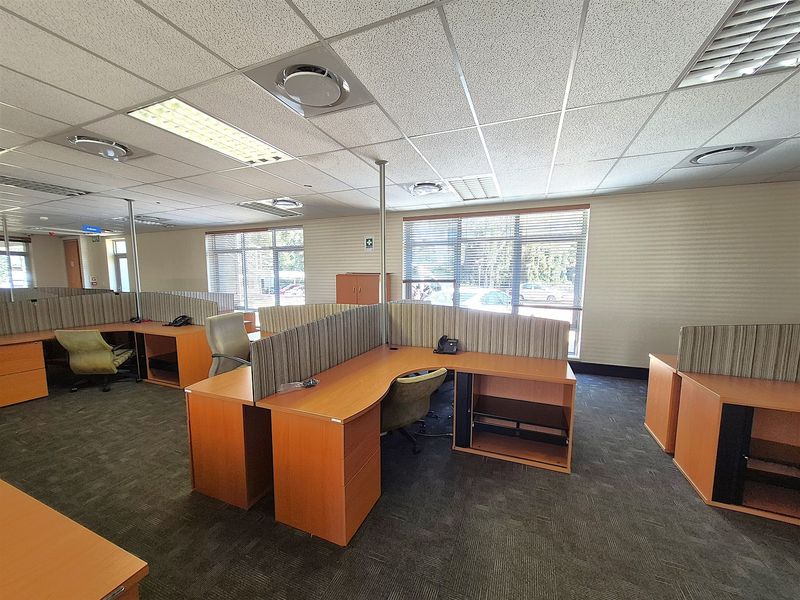 Open-plan office space featuring built-in workstations, suspended ceilings with integrated lighting, air conditioning vents, and large windows with venetian blinds.