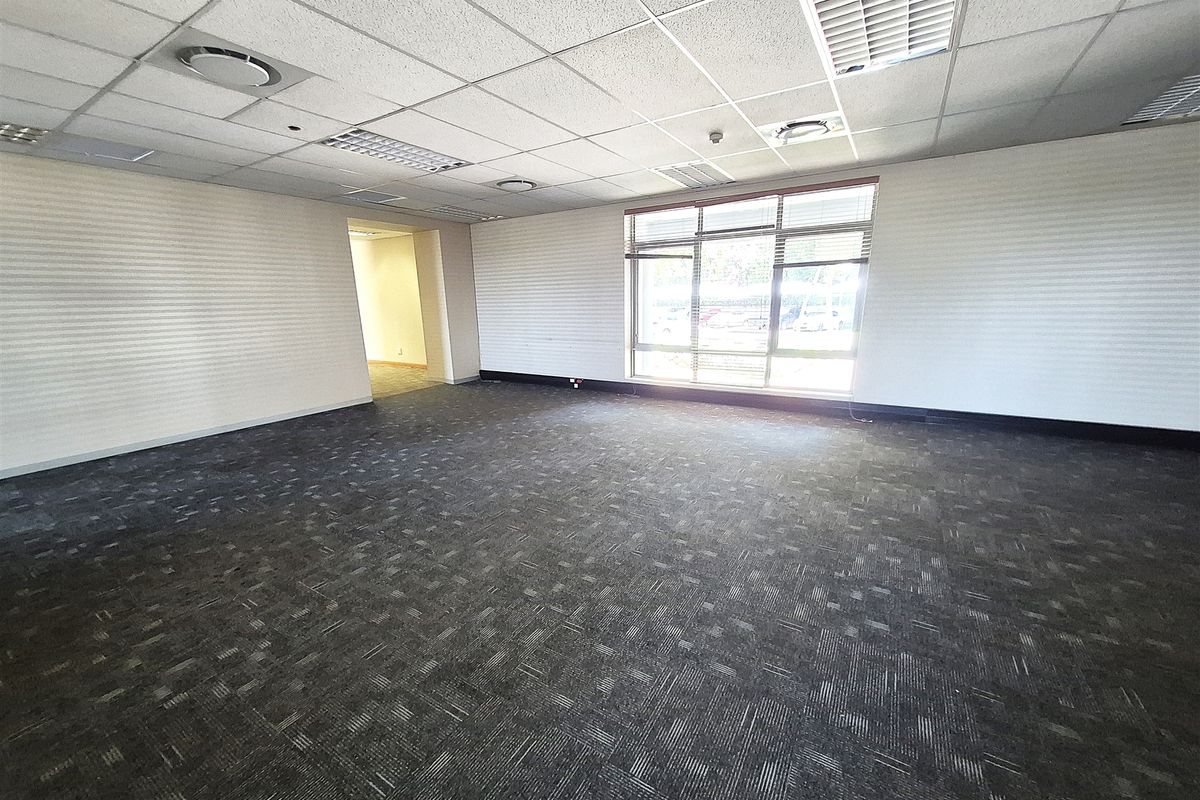 A spacious, empty office area featuring a suspended ceiling, recessed lighting, air conditioning vents, and large windows providing natural light.