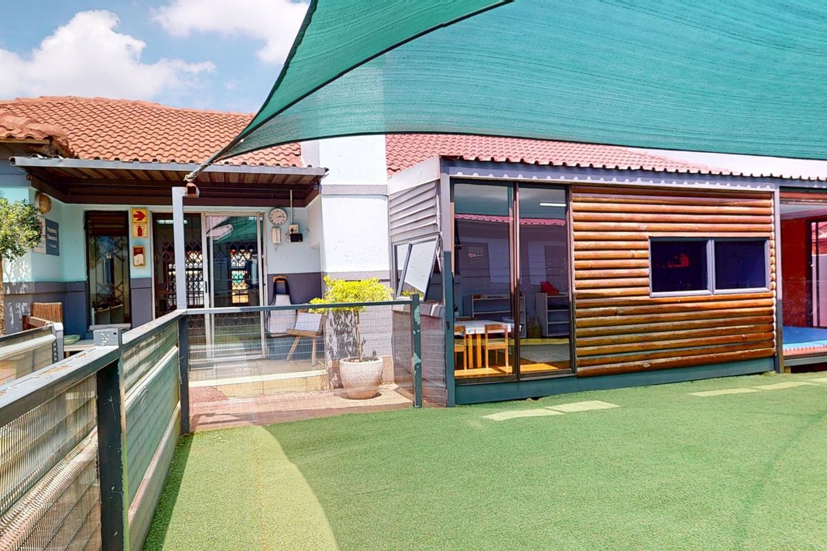 Rear exterior view of a commercial property featuring artificial turf, a large shade sail, and a modern log-style extension with sliding glass doors.