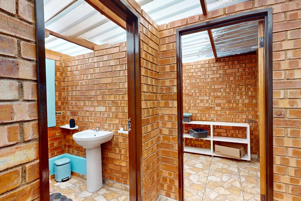 A functional outdoor-style bathroom featuring exposed brick walls, a pedestal basin, and a translucent roof, providing a rustic and practical utility space.