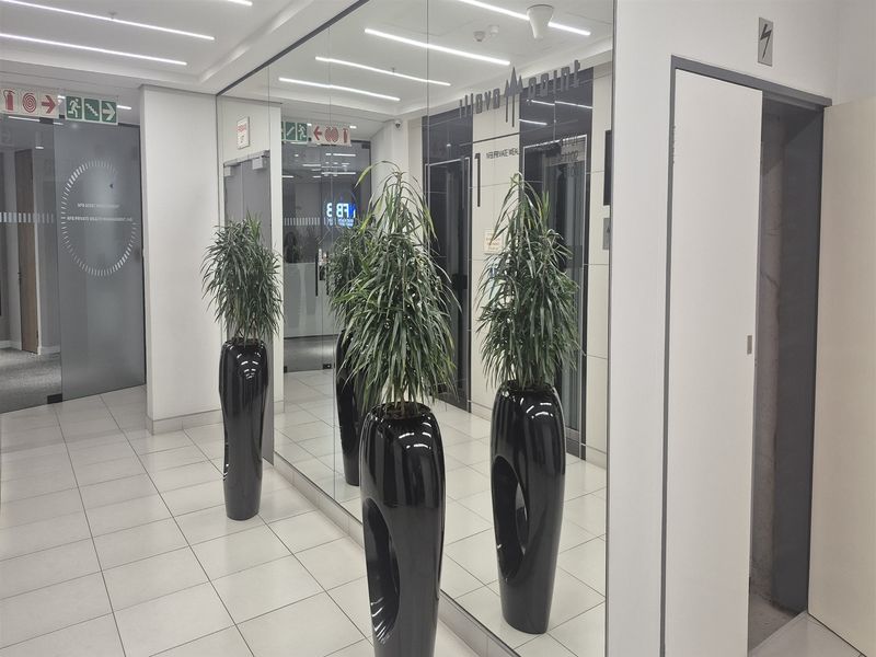 Modern office building hallway featuring tiled flooring, recessed linear ceiling lights, mirrored wall panels, and elevator access.
