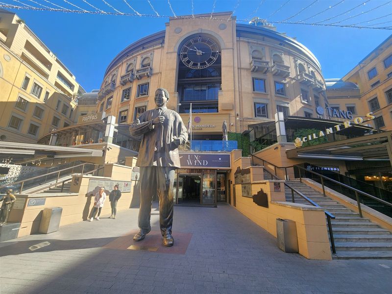 A wide-angle view of the Nelson Mandela Square plaza, featuring the iconic statue, clock tower, and surrounding commercial office and retail building architecture.