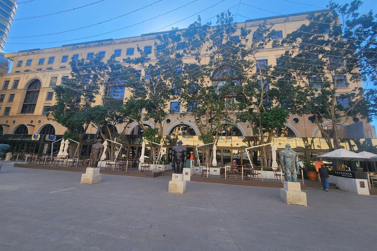 A wide-angle view of a multi-story office building facade featuring an expansive paved plaza, mature trees, and an integrated outdoor dining deck.