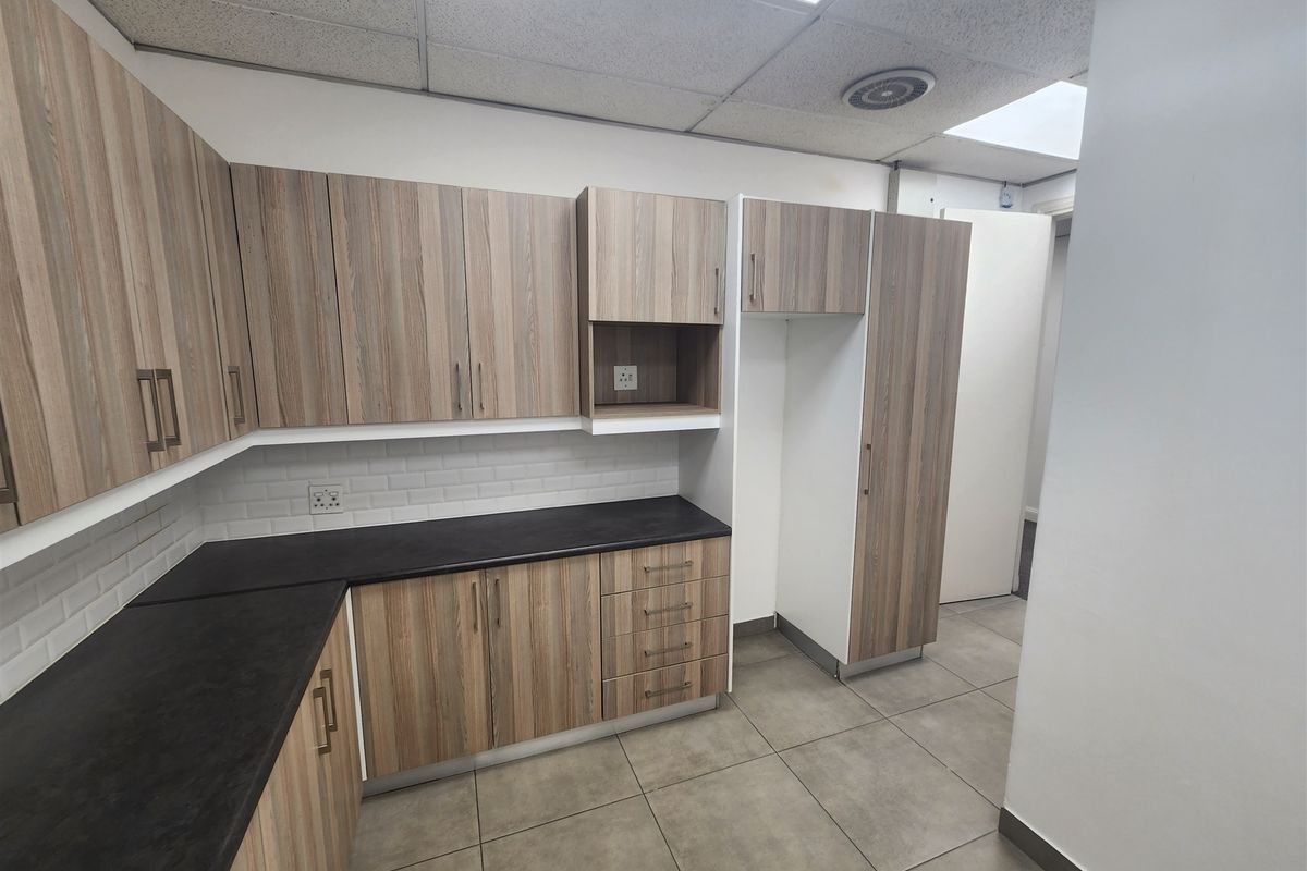A clean and modern office kitchenette featuring wood-look cabinetry, dark countertops, subway tile backsplash, and durable ceramic floor tiles.
