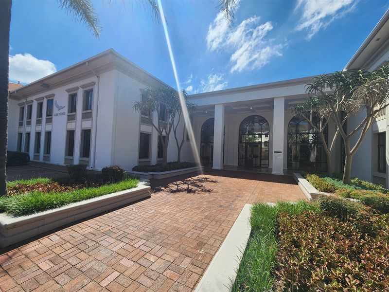 A well-maintained office building entrance featuring a brick-paved walkway, landscaped garden beds, and an elegant covered portico with arched glass doors.