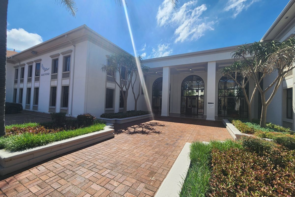A well-maintained office building entrance featuring a brick-paved walkway, landscaped garden beds, and an elegant covered portico with arched glass doors.