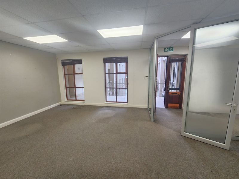 An empty office space featuring a suspended ceiling, recessed lighting, carpeted flooring, and glass partition doors with views into the building atrium.