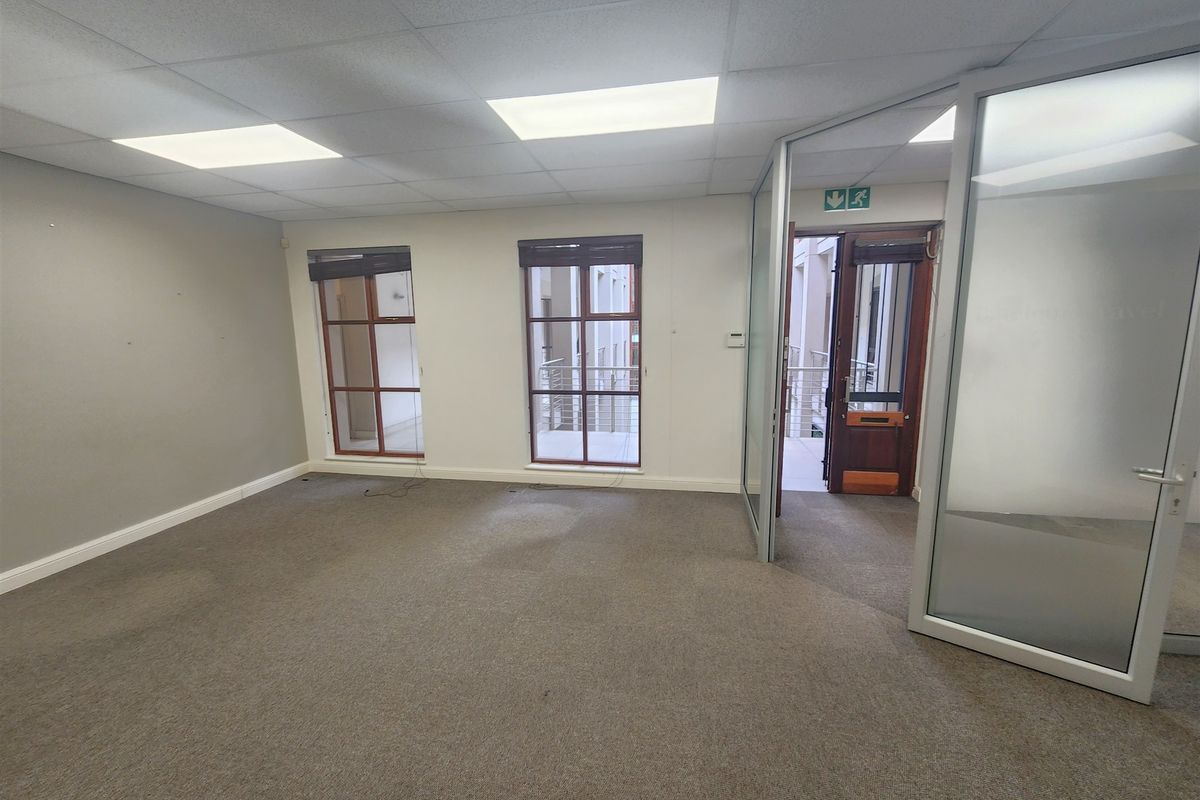 An empty office space featuring a suspended ceiling, recessed lighting, carpeted flooring, and glass partition doors with views into the building atrium.