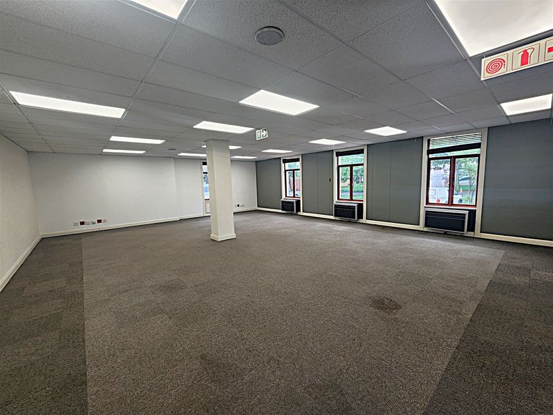 A spacious, empty open-plan office area featuring suspended ceilings, recessed lighting, carpeted flooring, and integrated window air conditioning units.