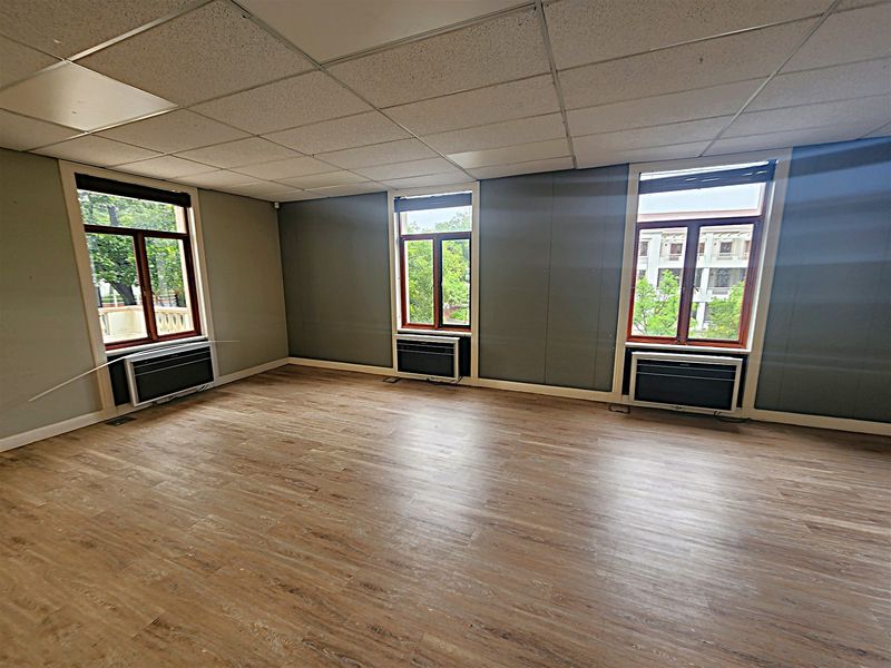 A spacious, empty office area featuring laminate flooring, a suspended ceiling, and three wall-mounted air conditioning units with natural light from three windows.
