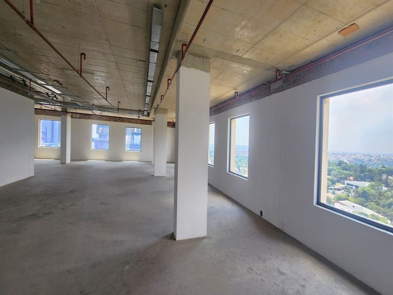 An expansive, open-plan office space featuring exposed concrete ceilings, structural columns, and large windows offering panoramic views, currently in a shell condition.