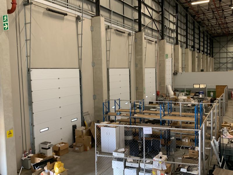 Spacious industrial warehouse interior featuring high ceilings with exposed steel trusses, multiple roller shutter doors, and concrete structural columns.