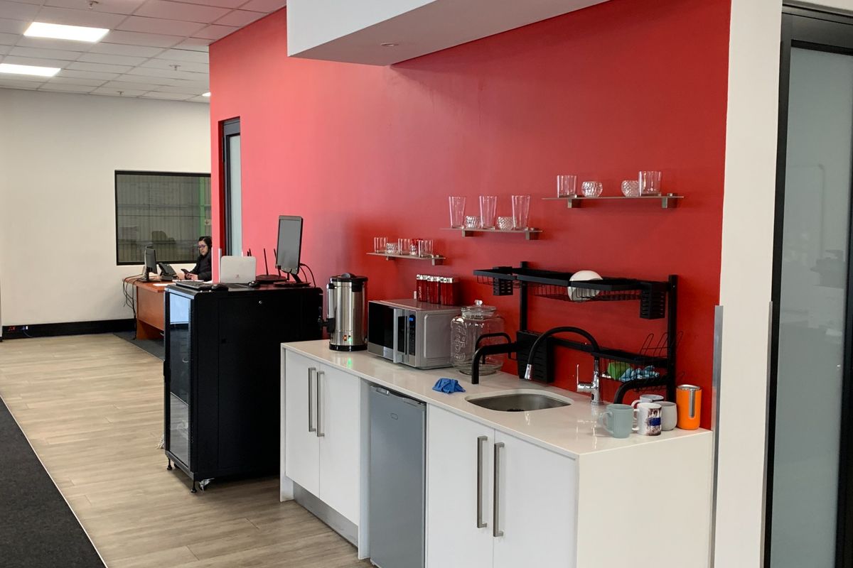 A compact kitchenette area featuring white cabinetry, a stainless steel sink, and a bold red accent wall within an office environment.