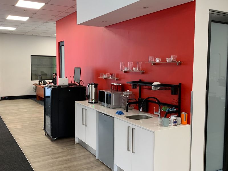 A compact kitchenette area featuring white cabinetry, a stainless steel sink, and a bold red accent wall within an office environment.