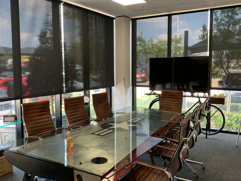 A modern office meeting room featuring floor-to-ceiling windows with roller blinds, recessed ceiling lighting, and carpeted flooring.