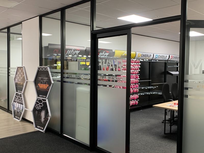 Modern industrial office space featuring glass partition walls, black aluminum framing, recessed lighting, and integrated retail shelving units.