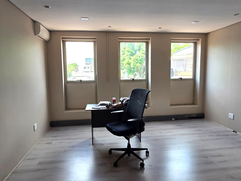 A bright office space featuring laminate flooring, recessed downlighting, and a wall-mounted air conditioning unit with three large windows providing natural light.