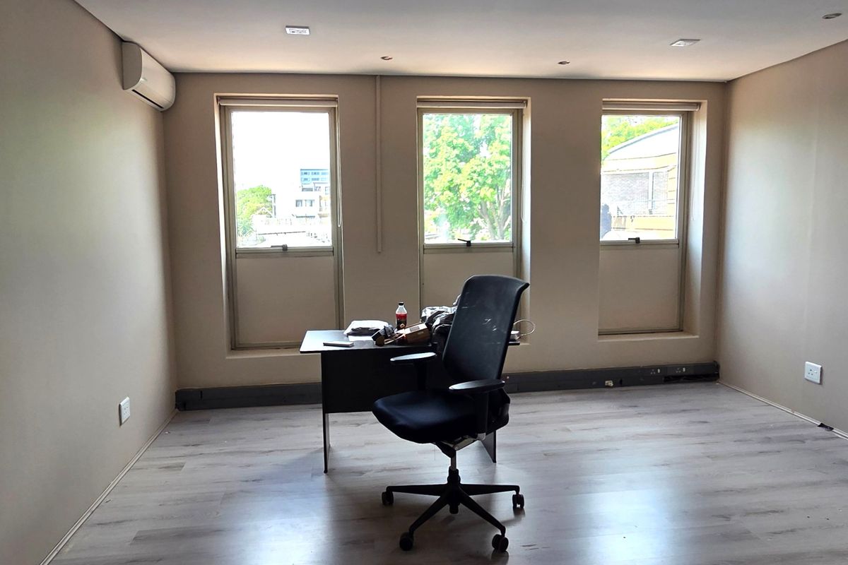 A bright office space featuring laminate flooring, recessed downlighting, and a wall-mounted air conditioning unit with three large windows providing natural light.