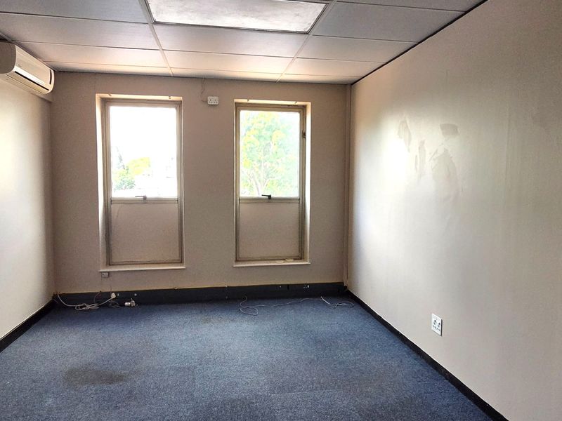 An empty office space featuring a suspended ceiling, split-system air conditioning, carpeted flooring, and dual windows providing natural light.
