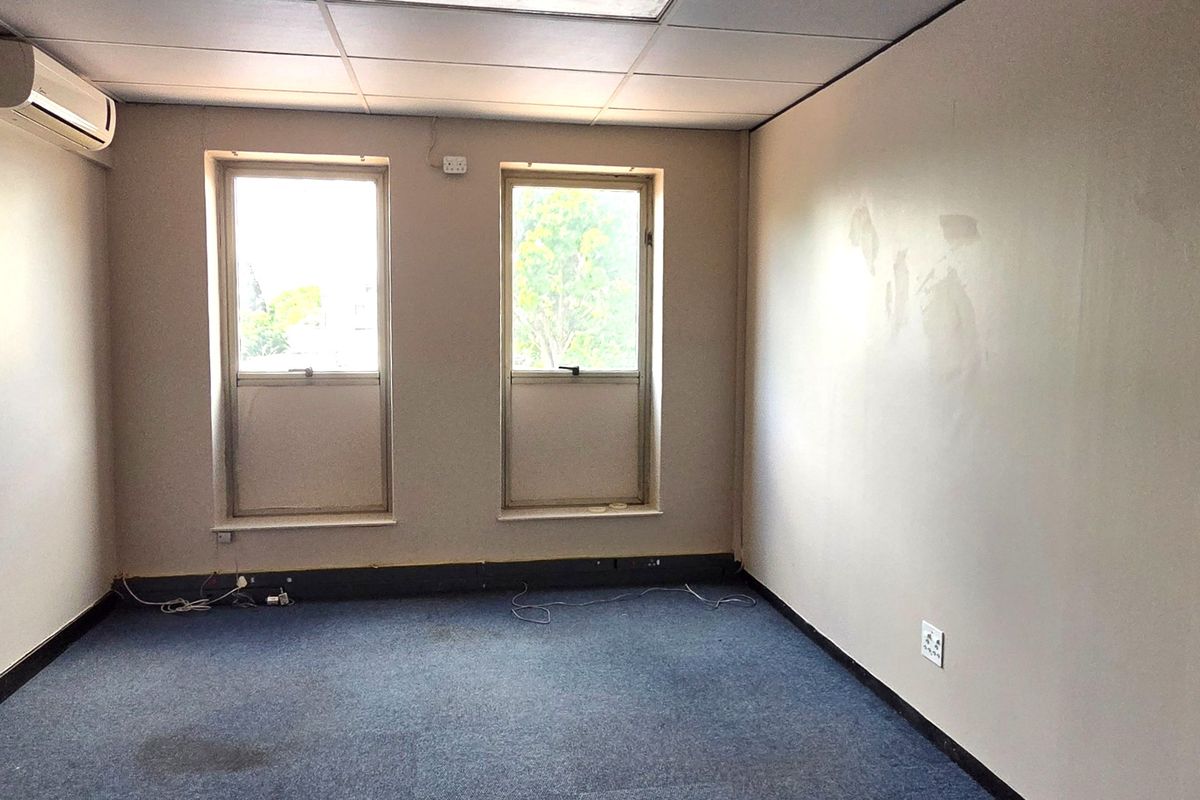 An empty office space featuring a suspended ceiling, split-system air conditioning, carpeted flooring, and dual windows providing natural light.