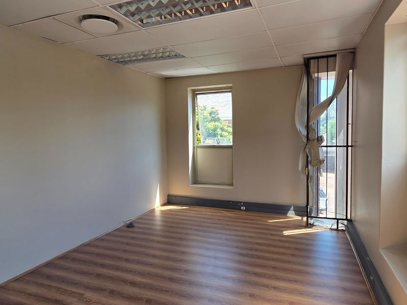 A vacant office space featuring laminate flooring, a suspended ceiling with recessed lighting, and security bars on the window.