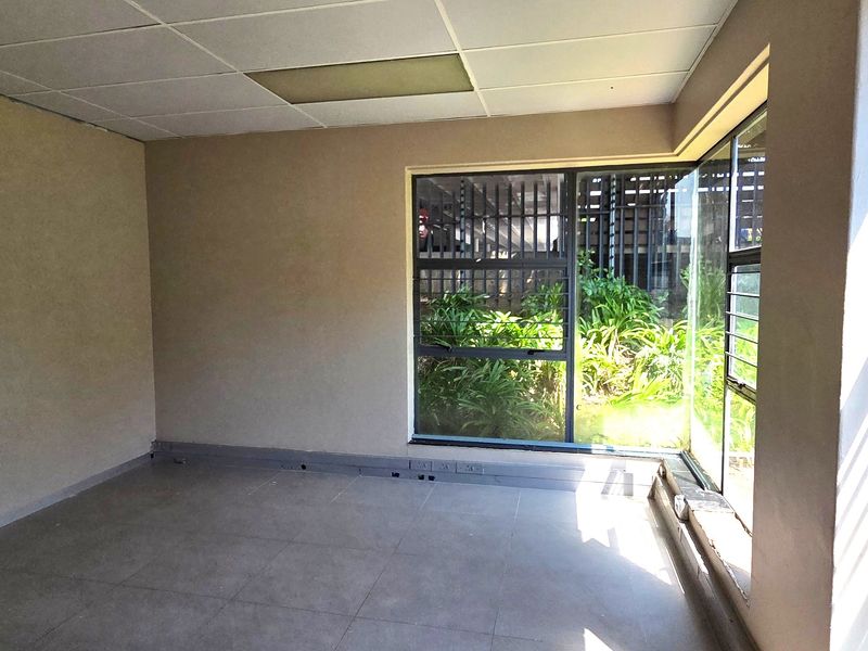 An empty office space featuring tiled flooring, a suspended ceiling, and a corner window with security bars overlooking a garden area.