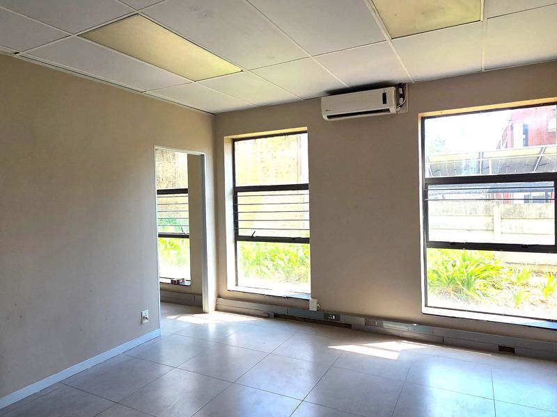 A vacant office space featuring tiled flooring, a suspended ceiling, and a wall-mounted split-system air conditioning unit with natural light from large windows.